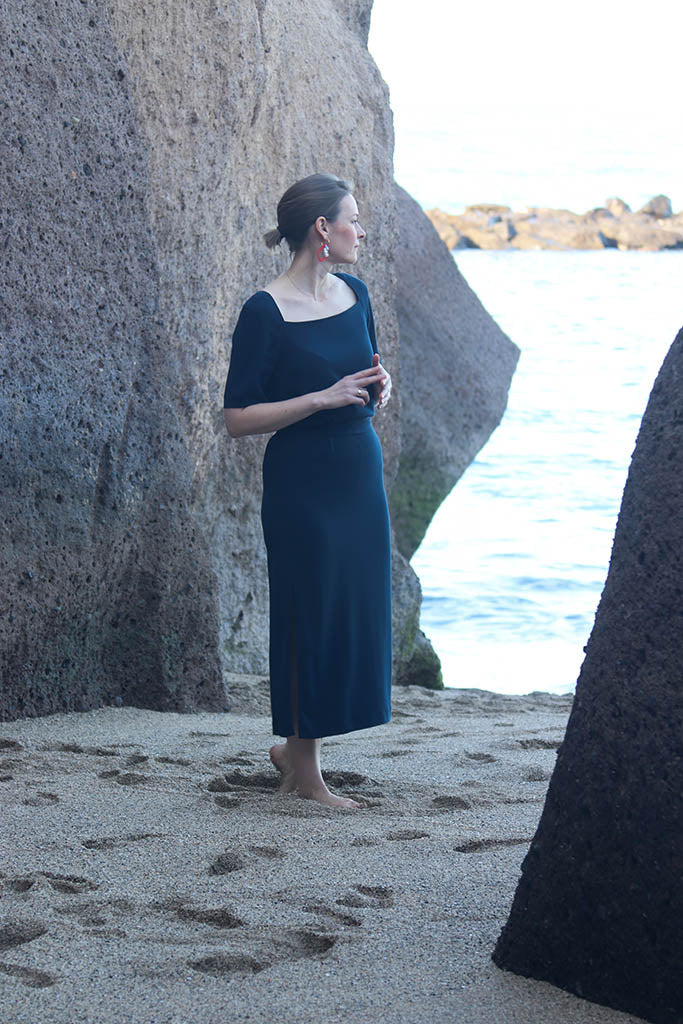 Frau in dunkelblauem langem Kleid, barfuß am Strand, rote Ohrringe