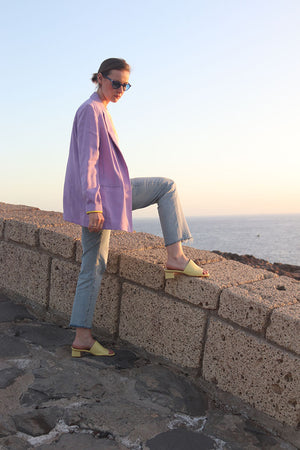 Frau in heller Jeans und Flieder Blazer vor Strandmauer stehend