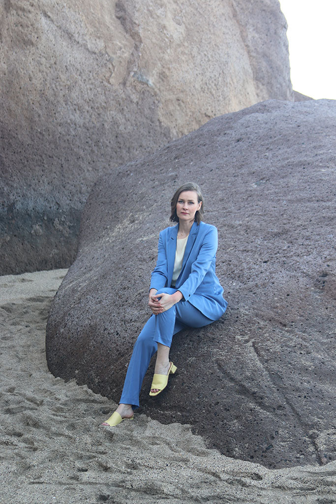Frau in Kornblauem Anzug und heller Seidenbluse auf Felsen sitzend