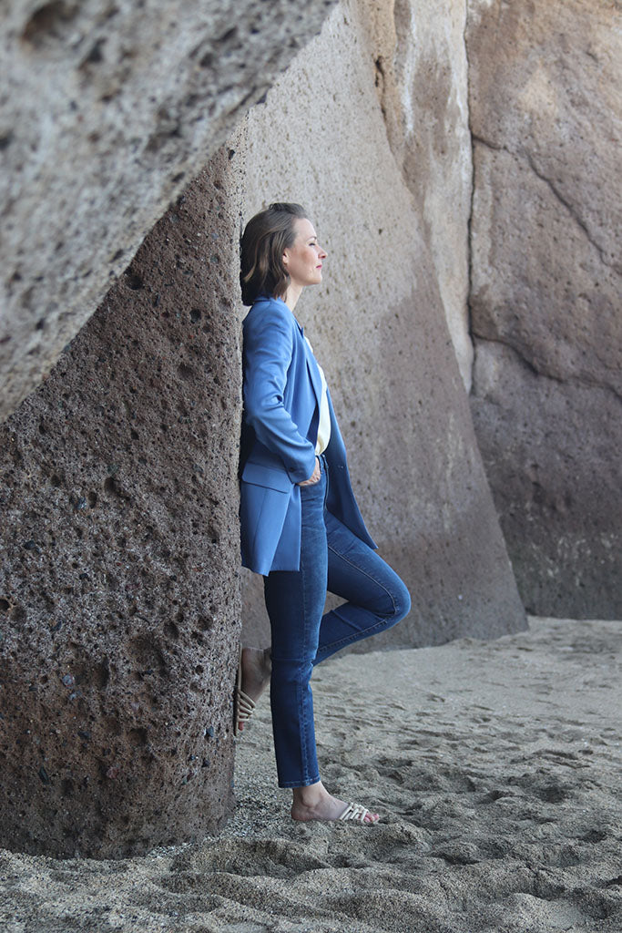 Frau in schmaler Jeans und hellblauem Blazer steht am Strand gelehnt an Felswand
