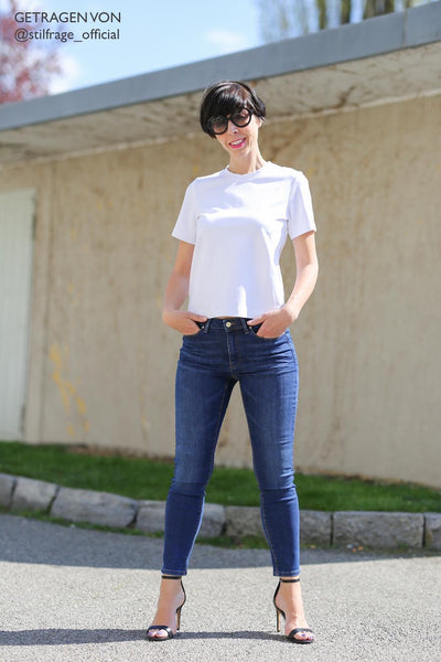 Frau in schmaler Jeans, weißem T-Shirt und schwarzen hohen Riemchensandalen steht auf Straße